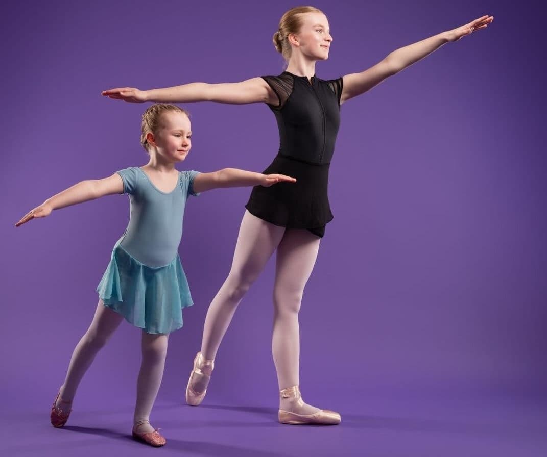 A teacher and young pupil holding matching arabesques on a purple backdrop