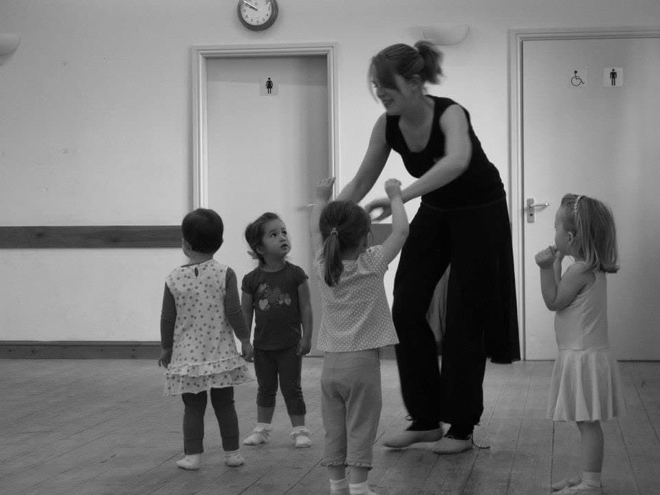 Black and white photo of Jess leading an early baby ballet class in a hall