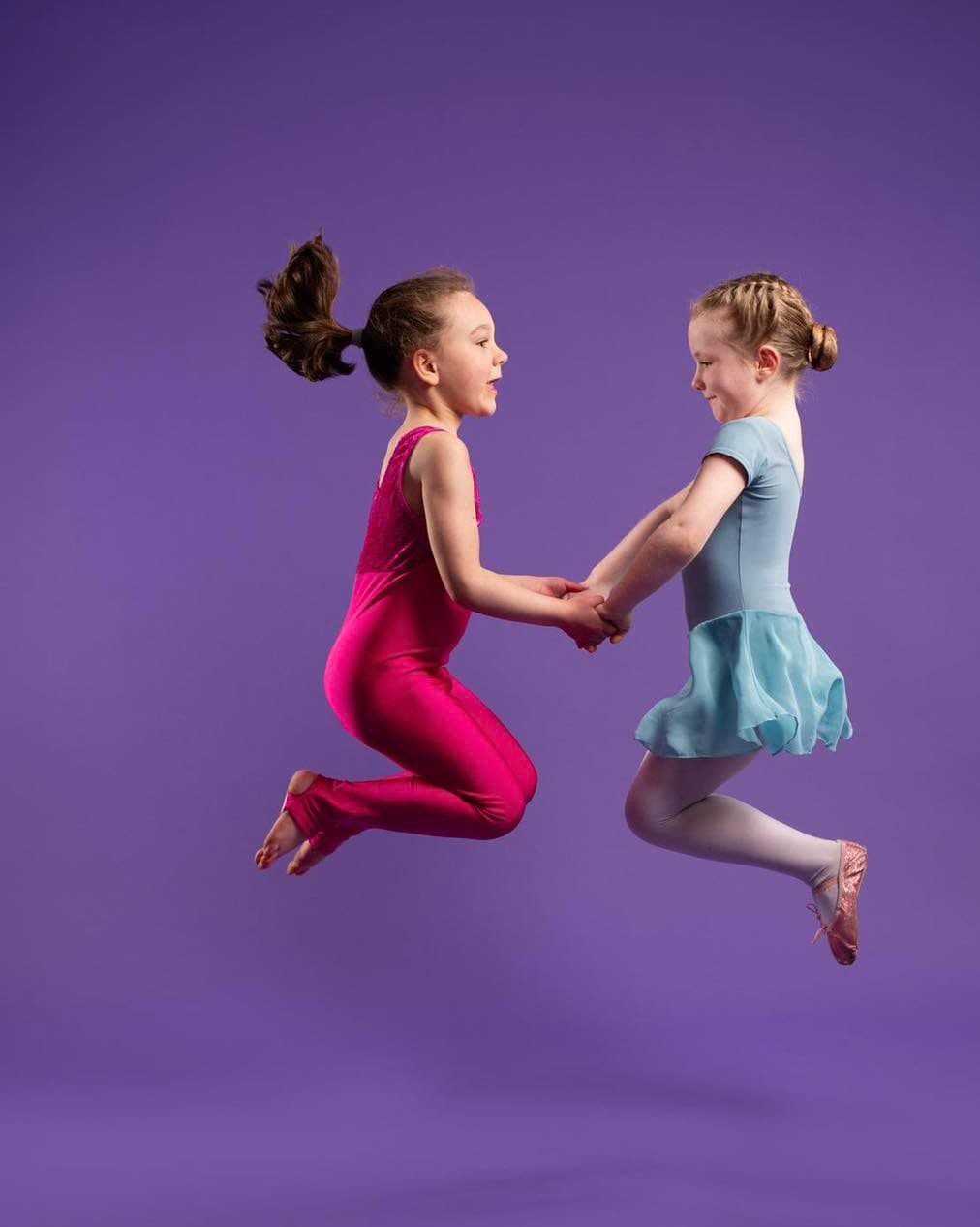 Two young Jazzy J's dancers leaping hand in hand in pink and pale blue