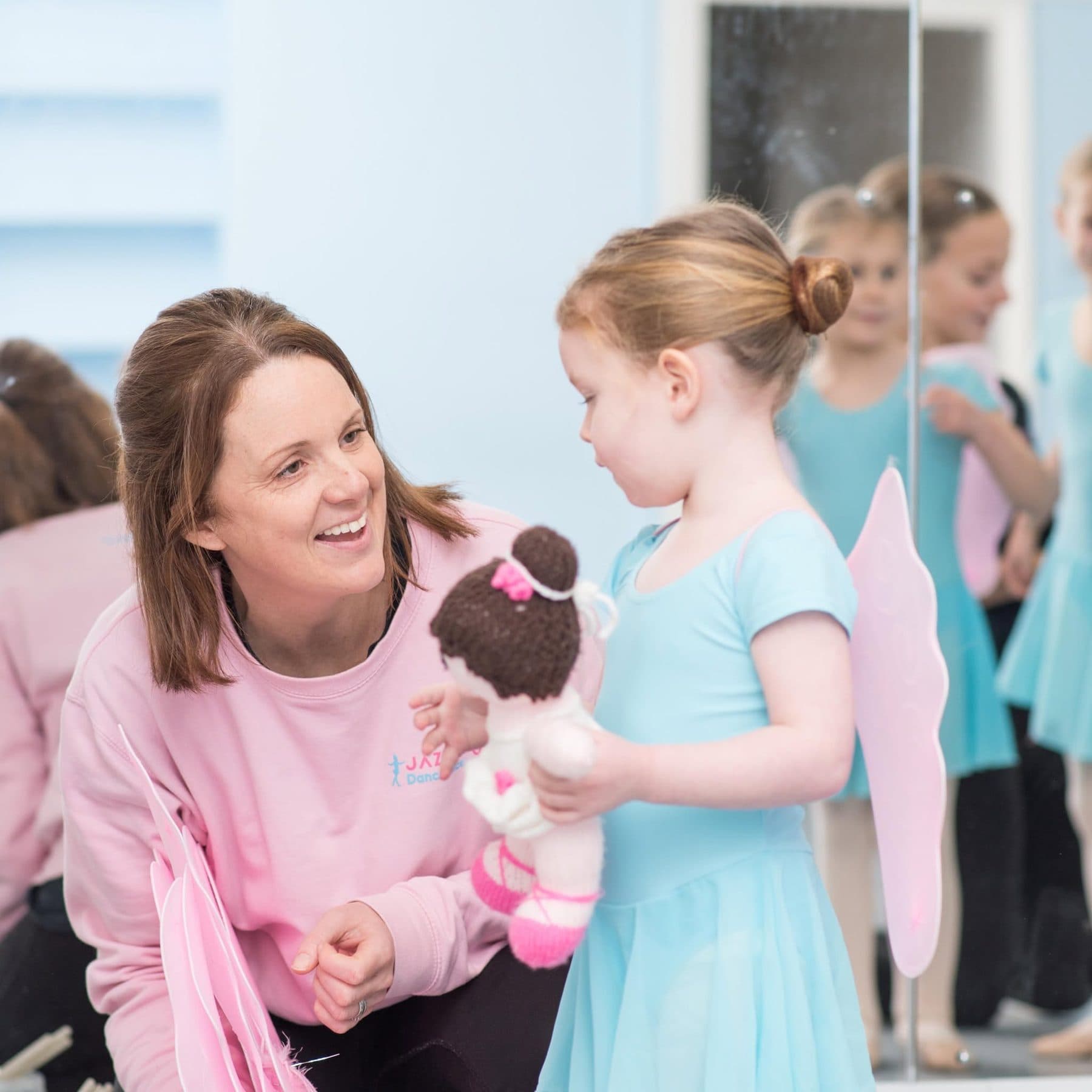 Jess, our Principal, kneeling to chat with a young ballet pupil holding a knitted ballerina doll